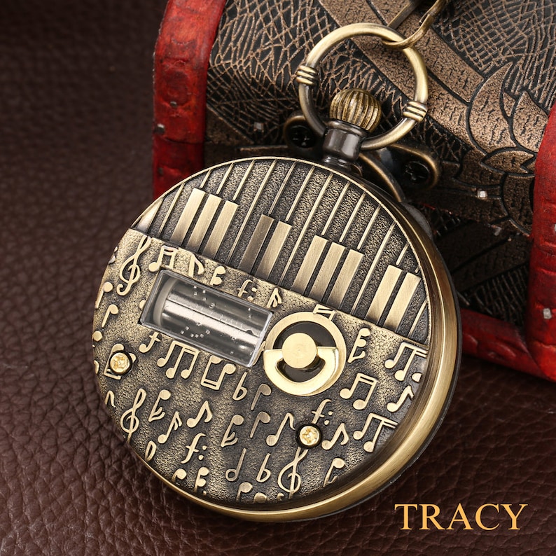 May include: A gold-toned pocket watch with a musical theme. The watch face features a piano keyboard design and musical notes. The watch has a clear window revealing a small, intricate mechanism.
