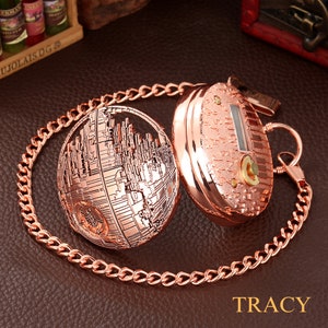May include: A rose gold pocket watch with a chain. The watch face is a detailed design of the Death Star from Star Wars. The back of the watch is plain with a small keyhole.