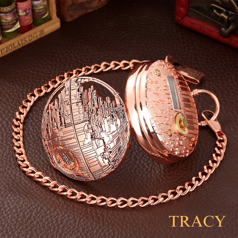 May include: A rose gold pocket watch with a chain. The watch face is a detailed design of the Death Star from Star Wars. The back of the watch is plain with a small keyhole.