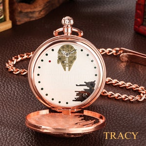 May include: A rose gold pocket watch with a white face and a detailed image of the Millennium Falcon from Star Wars. The watch has a chain attached to it.