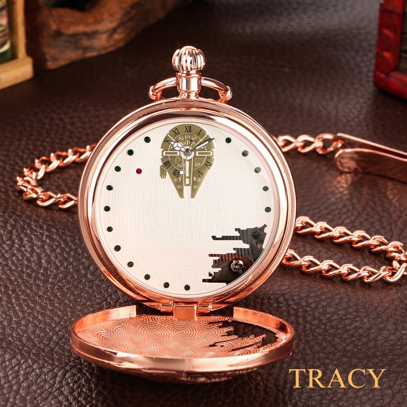 May include: A rose gold pocket watch with a white face and a detailed image of the Millennium Falcon from Star Wars. The watch has a chain attached to it.