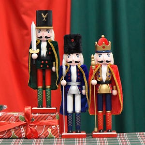 Op de afbeelding: Drie houten notenkrakers. De notenkraker links draagt een groen en rood pak met gouden versieringen en houdt een zwaard vast. De notenkraker in het midden draagt een blauw en wit pak met gouden versieringen en houdt een speer vast. De notenkraker rechts draagt een rood en gouden pak met gouden versieringen en houdt een staf vast.