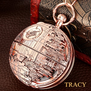 May include: A rose gold pocket watch with a detailed design of the Death Star from Star Wars. The watch has a clear face and a chain attached.