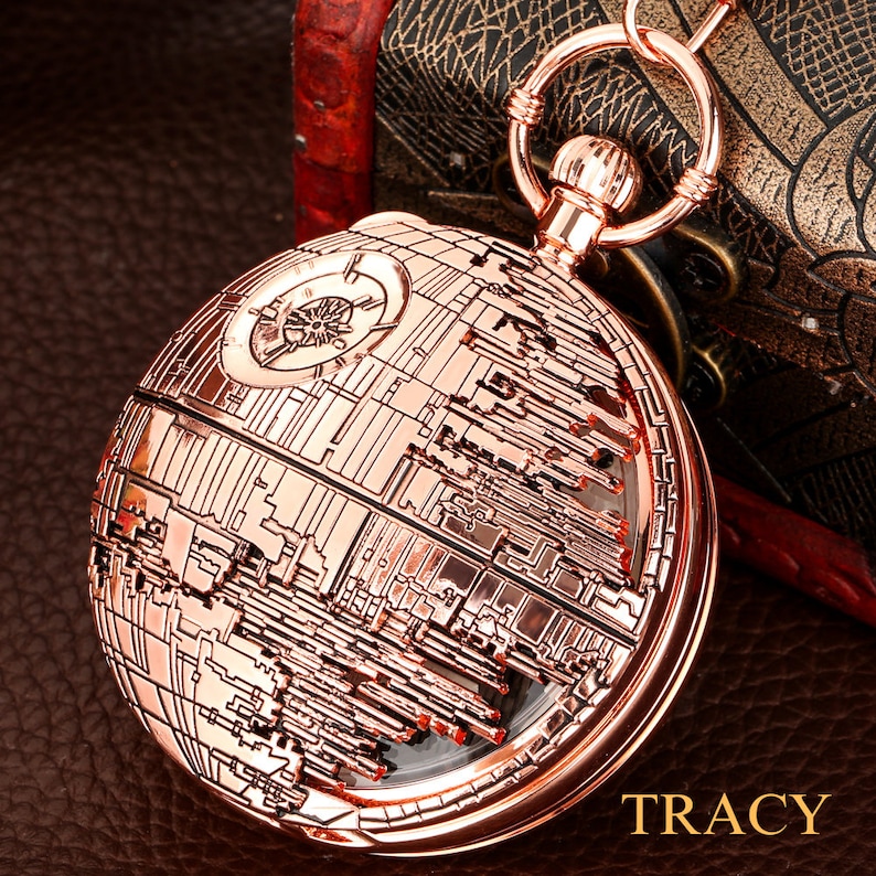 May include: A rose gold pocket watch with a detailed design of the Death Star from Star Wars. The watch has a clear face and a chain attached.