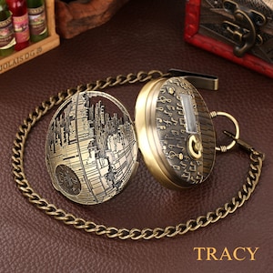 May include: A gold-toned pocket watch with a chain. The watch face is covered with musical notes and has a small window for the time. The back of the watch is a detailed design of the Death Star from Star Wars.