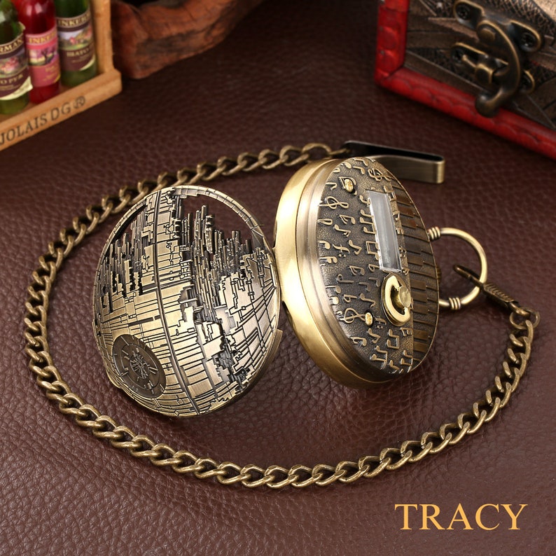 May include: A gold-toned pocket watch with a chain. The watch face is covered with musical notes and has a small window for the time. The back of the watch is a detailed design of the Death Star from Star Wars.