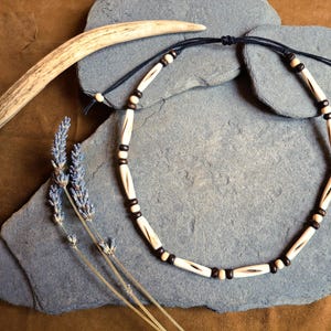 Può includere: Una collana di perline in osso bianco e marrone con un cordoncino nero. La collana è su una superficie di pietra grigia con un pezzo di corna di cervo e fiori di lavanda.