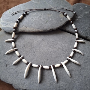 May include: A handcrafted necklace featuring off-white, elongated pendant beads interspersed with dark brown and white round beads. The necklace is strung on a brown cord, creating a rustic and natural aesthetic. The necklace is displayed on a gray stone surface.
