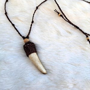 Può includere: Una collana artigianale con un ciondolo a forma di corno color crema. Il ciondolo è racchiuso in una manica lavorata a maglia marrone scuro e sospeso a un cordoncino marrone abbinato. Il cordoncino è adornato con piccole perline di legno.