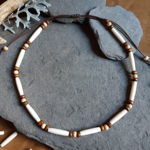 May include: A brown leather cord necklace with white and brown cylindrical beads. The necklace is lying on a gray stone surface.