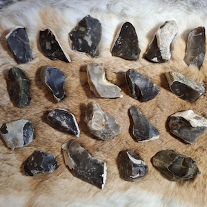 Puede incluir: Una colección de diversas herramientas de pedernal gris oscuro y negro con bordes blancos y beige, dispuestas sobre una superficie similar a piel marrón y beige. Las herramientas varían en forma y tamaño, mostrando una estética primitiva.