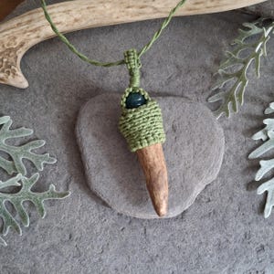 Può includere: Una collana fatta a mano con un ciondolo in corno avvolto in macramè verde. Una perla verde scuro adorna il ciondolo, sospeso a un cordino verde. La collana è appoggiata su una superficie di pietra grigia.