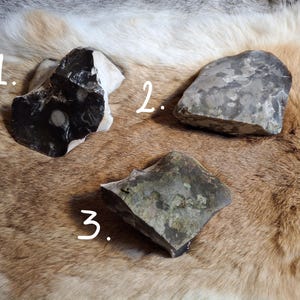 Puede incluir: Tres piedras de forma irregular con diferentes colores y texturas se muestran sobre una superficie similar a la piel. Una piedra es principalmente negra y blanca, otra gris y la tercera es una mezcla de gris y verde.