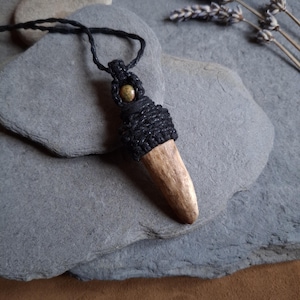 May include: A handcrafted pendant necklace with a natural antler pendant wrapped in black cord. The antler is a light brown colour, and the necklace has a black cord. A small, round, olive-green bead is at the top of the pendant.