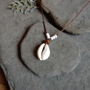 May include: A brown cord necklace with white beads and a cowrie shell pendant. The necklace is displayed on a grey stone surface.