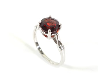 Puede incluir: Un anillo de plata con una piedra preciosa roja engastada en garras. El anillo tiene un diseño delicado con una pequeña piedra de acento en el lateral.
