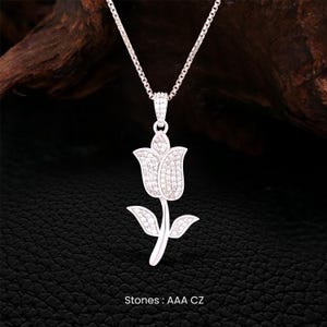 May include: A silver-colored pendant necklace featuring a tulip design. The tulip pendant is encrusted with small, sparkling stones. The necklace hangs on a delicate chain. The text "Stones: AAA CZ" is visible at the bottom of the image.