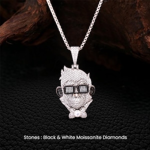 May include: A silver-toned pendant necklace featuring a stylized face with sunglasses, encrusted with white and black moissanite diamonds. The pendant hangs from a delicate chain. The text at the bottom reads: Stones: Black & White Moissanite Diamonds.