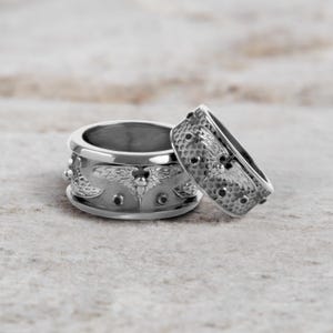 May include: Two silver rings with intricate designs. One ring is wider, featuring an embossed pattern of wings and small black accents. The other ring is narrower, with a similar design, textured surface, and black accents. Both rings are on a neutral surface.