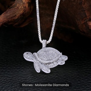 Könnte beinhalten: Eine silberne Kettenkette mit einem Schildkrötenanhänger. Die Schildkröte ist mit klaren, funkelnden Steinen bedeckt. Der Text "Stones: Moissanite Diamonds" befindet sich unter der Schildkröte.