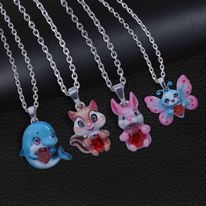 Könnte beinhalten: Vier Halsketten mit silbernen Ketten und Anhängern. Die Anhänger sind in Form eines blauen Delfins, eines braunen Eichhörnchens, eines rosa Kaninchens und eines blauen Schmetterlings gestaltet. Jeder Anhänger hat einen roten herzförmigen Edelstein.