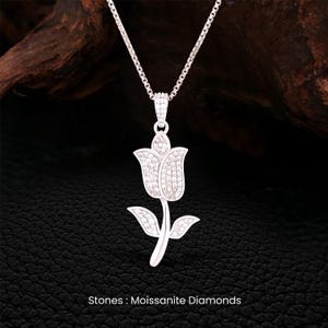 May include: A silver-toned pendant necklace featuring a tulip design. The pendant is encrusted with small, sparkling stones, and hangs from a delicate chain. The text at the bottom of the image reads "Stones: Moissanite Diamonds."