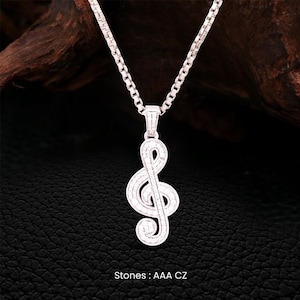 Pode incluir: Um colar prateado com um pendente de clave de sol adornado com pequenas pedras brilhantes. A corrente é do tipo elo quadrado. O texto "Stones: AAA CZ" está na parte inferior da imagem.