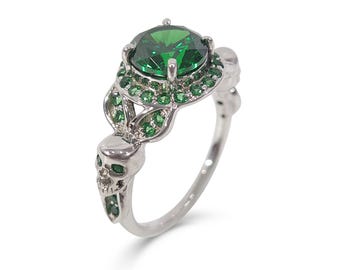 Könnte beinhalten: Ein silberner Ring mit einem großen grünen Smaragd, der von kleineren grünen Edelsteinen umgeben ist. Der Ring hat zwei Totenkopf-Designs auf der Band.