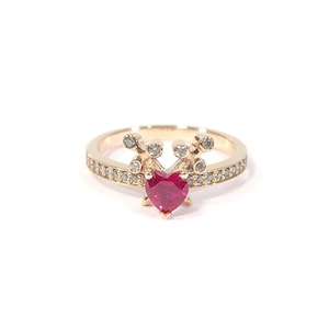 Puede incluir: Un anillo de oro con una rubí roja en forma de corazón y una banda de pequeños diamantes.