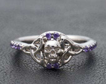 May include: A silver ring with a skull design and purple gemstones. The skull is surrounded by a Celtic knot design.