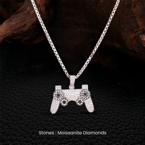 May include: A silver pendant necklace with a controller design. The controller is adorned with small, sparkling stones, with black and white accents on the buttons. The text "Stones: Moissanite Diamonds" is at the bottom of the image.