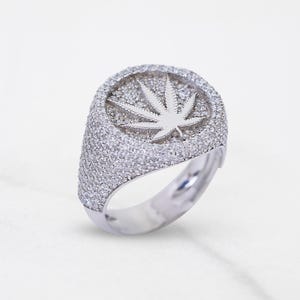 Puede incluir: Un anillo plateado con un diseño de hoja de cannabis. El anillo presenta una cara redonda con una hoja detallada, rodeada de pequeñas piedras brillantes. La banda es lisa y pulida.