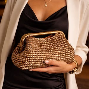 Puede incluir: Un clutch dorado tejido con un asa dorada. El clutch se sostiene frente a un vestido negro y una blazer blanca. También se ven un collar y una pulsera dorados.