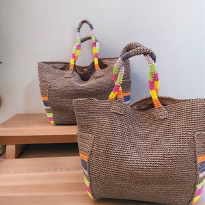 May include: Two brown woven tote bags with colorful striped accents and handles. The handles are braided with vibrant rainbow-colored yarn. The bags have a rectangular shape and are displayed on a wooden surface.