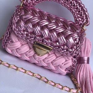 May include: A pink crocheted handbag with a gold chain strap and a pink tassel. The bag has a woven design and a gold clasp.