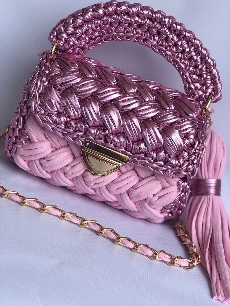 May include: A pink crocheted handbag with a gold chain strap and a pink tassel. The bag has a woven design and a gold clasp.