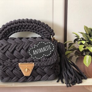 Peut inclure: Un sac &agrave; main en crochet gris fonc&eacute; avec un fermoir dor&eacute; et un long pompon. Le sac a un design tress&eacute; et l'&eacute;tiquette indique "Anthracite".