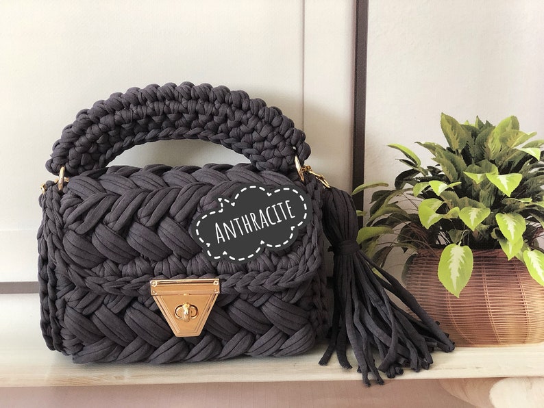 Peut inclure: Un sac &agrave; main en crochet gris fonc&eacute; avec un fermoir dor&eacute; et un long pompon. Le sac a un design tress&eacute; et l'&eacute;tiquette indique "Anthracite".