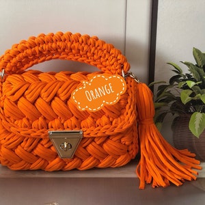 Peut inclure: Sac &agrave; main en crochet orange avec un design tress&eacute; et un fermoir dor&eacute;. Le sac a un long pompon orange.