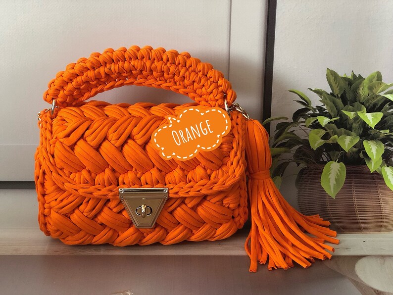 Peut inclure: Sac &agrave; main en crochet orange avec un design tress&eacute; et un fermoir dor&eacute;. Le sac a un long pompon orange.