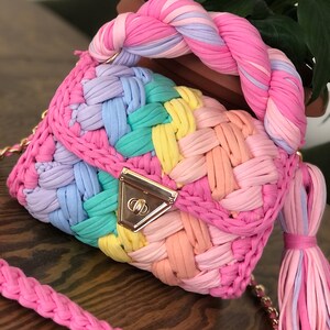 May include: A rainbow-colored crocheted handbag with a braided handle and a gold clasp. The bag has a matching shoulder strap.
