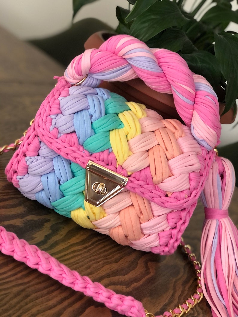 May include: A rainbow-colored crocheted handbag with a braided handle and a gold clasp. The bag has a matching shoulder strap.