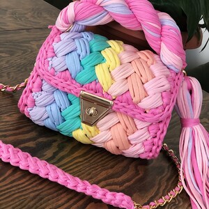 May include: A colorful crocheted handbag with a braided handle and a matching tassel. The bag is made of a variety of pastel colors, including pink, blue, green, and yellow.