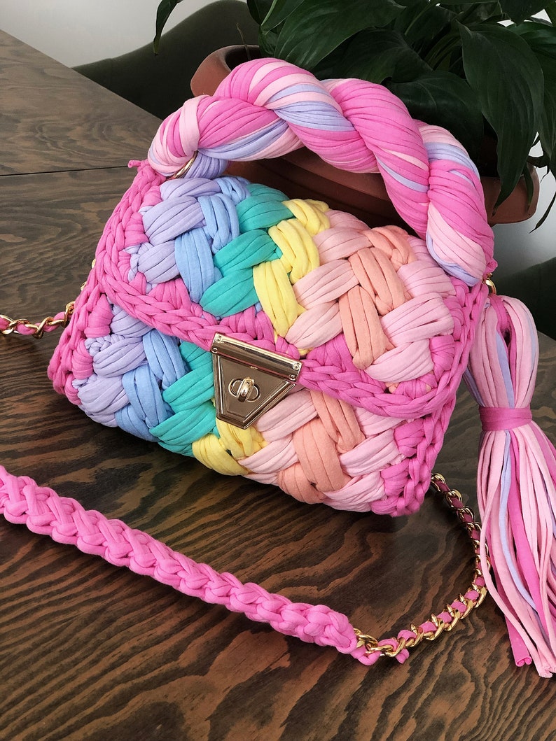 May include: A colorful crocheted handbag with a braided handle and a matching tassel. The bag is made of a variety of pastel colors, including pink, blue, green, and yellow.