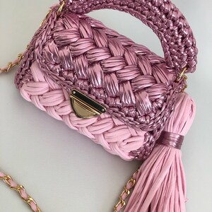 May include: A pink crocheted handbag with a gold chain strap and a tassel. The bag has a woven design and a top handle.
