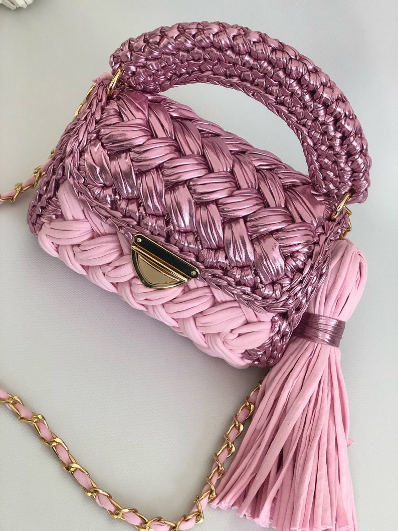 May include: A pink crocheted handbag with a gold chain strap and a tassel. The bag has a woven design and a top handle.