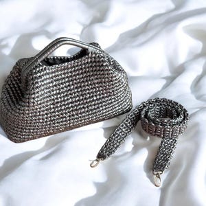 May include: A silver-toned handbag with a matching shoulder strap. The bag has a woven texture and a small handle. The strap is detachable with silver-colored clasps. The bag and strap are set against a white background.