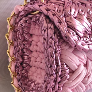 May include: A pink crocheted handbag with a gold chain strap. The bag is made of a combination of smooth and textured fabric.