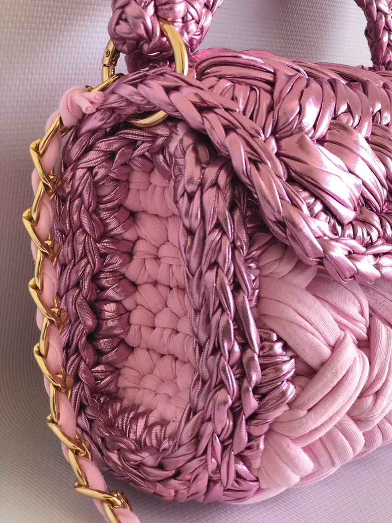 May include: A pink crocheted handbag with a gold chain strap. The bag is made of a combination of smooth and textured fabric.