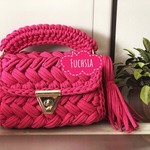 Peut inclure: Un sac &agrave; main en crochet fuchsia vif avec un fermoir dor&eacute; et un pompon. Le sac a une texture tiss&eacute;e et est &eacute;tiquet&eacute; "Fuchsia".